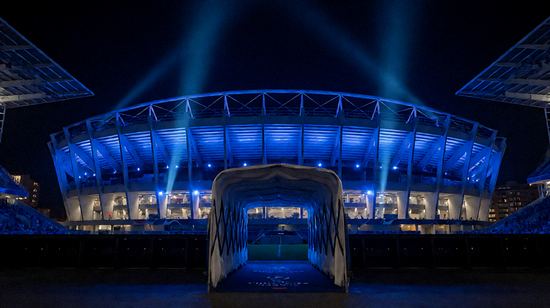 欧冠联赛比赛日夜晚的球场外观，蓝色的灯光笼罩整个体育场，球员通道入口清晰可见，营造出顶级赛事的庄严氛围